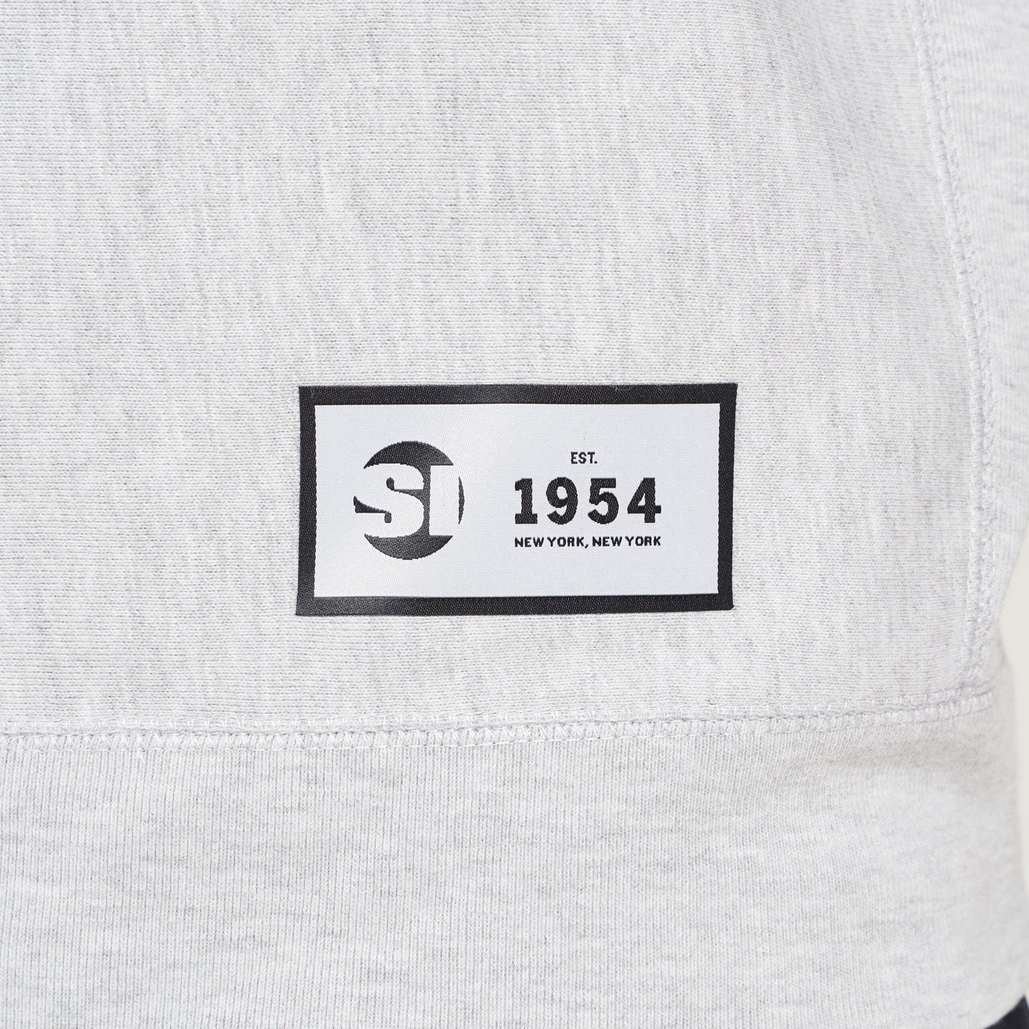 A label on a light gray sweatshirt featuring a black rectangular patch with the text SI EST 1954 NEW YORK NEW YORK