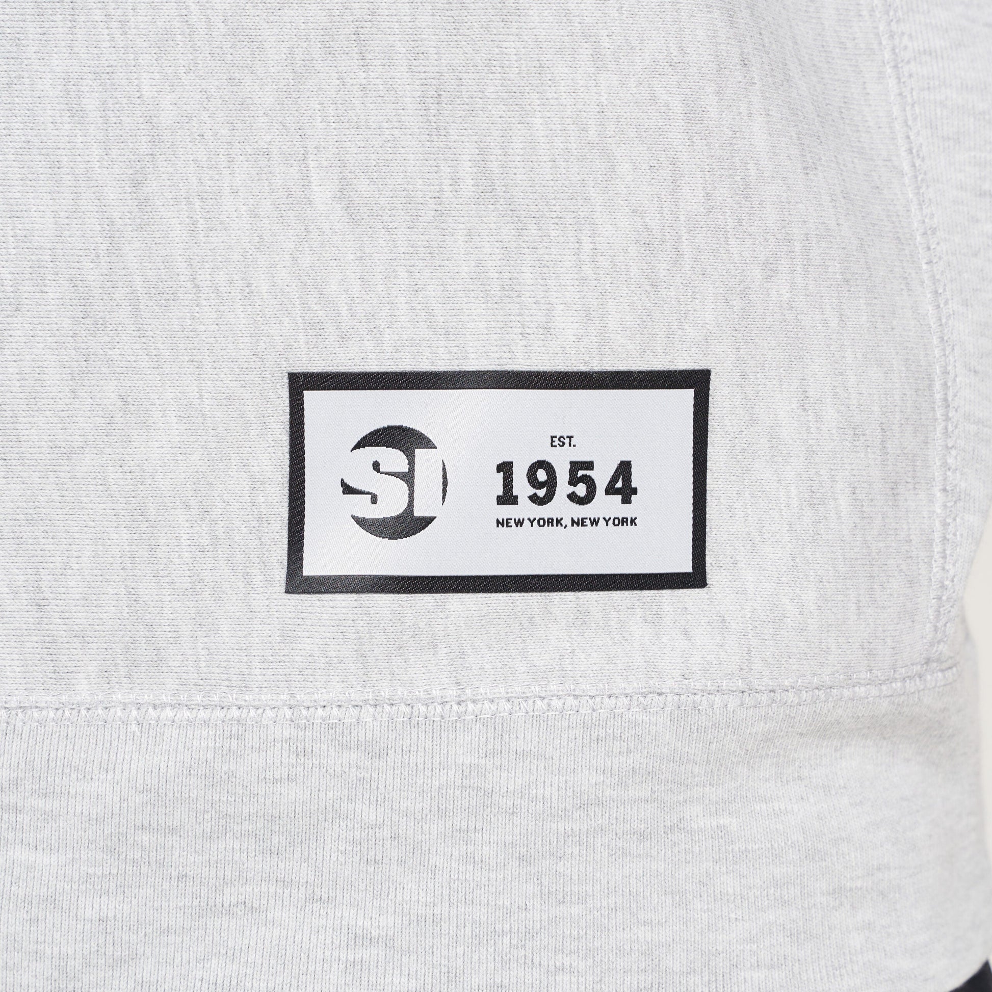 A label on a light gray sweatshirt featuring a black rectangular patch with the text SI EST 1954 NEW YORK NEW YORK