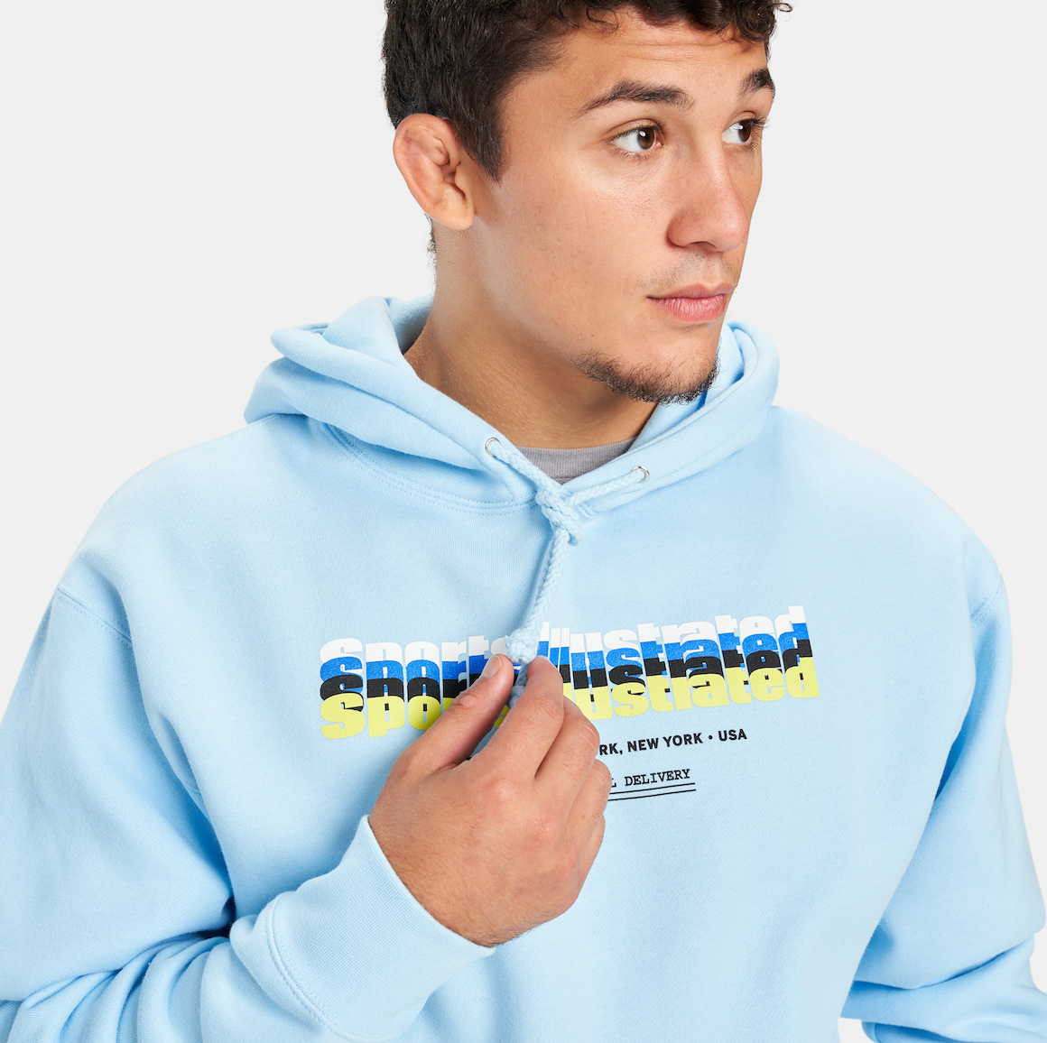 A young man wearing a light blue hoodie with Sports Illustrated printed on it posing thoughtfully while touching the drawstring