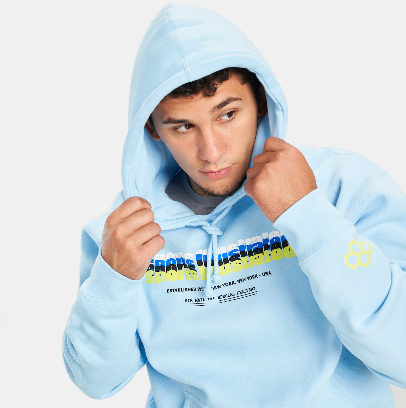 A young man wearing a light blue hoodie with bold graphic text and a playful design adjusting the hood of his sweatshirt