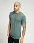 A man with a shaved head and a prominent beard is wearing a fitted green t-shirt showcasing his colorful tattoos on his arm against a neutral background