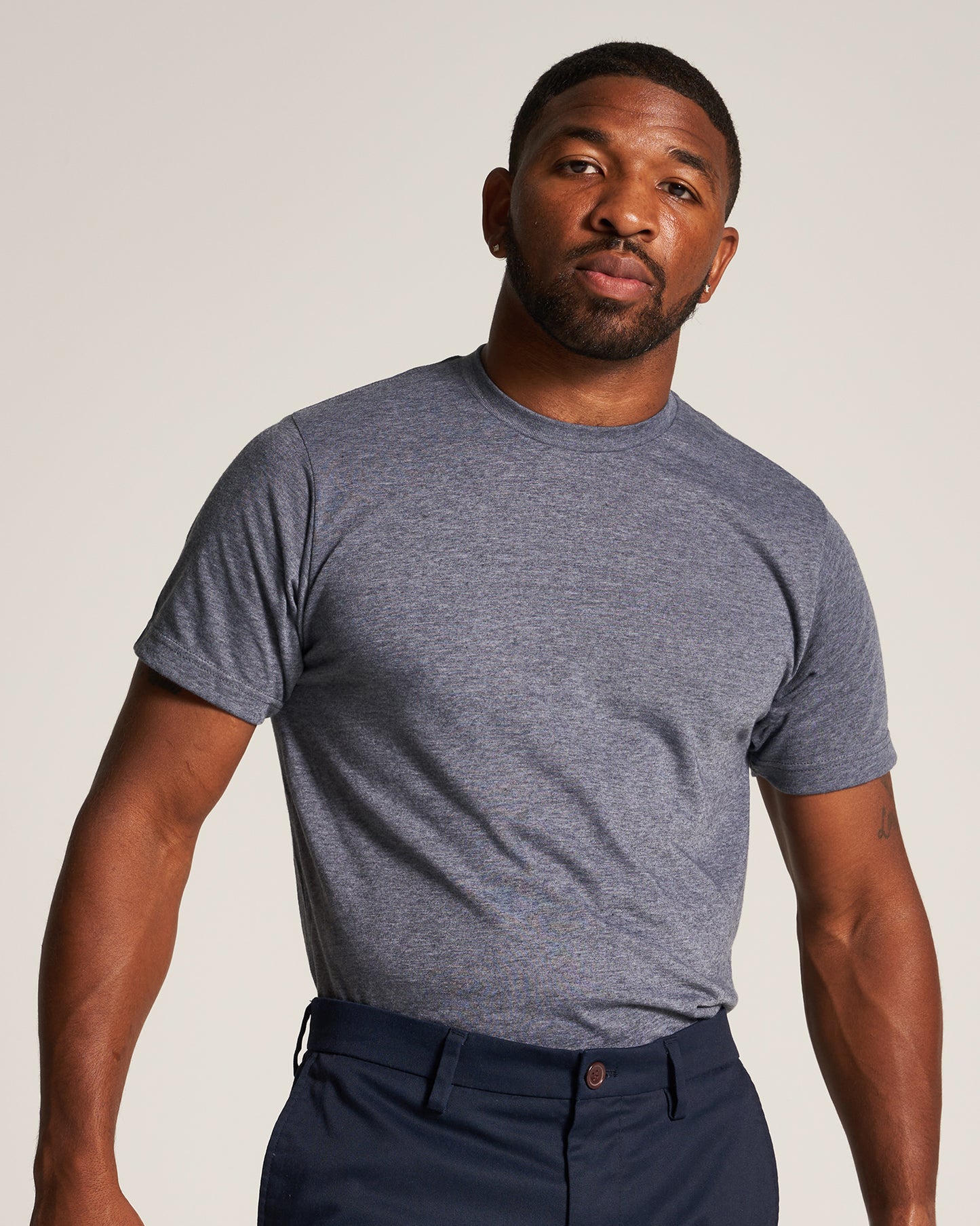 A confident man poses in a fitted gray t-shirt and navy blue pants showcasing a casual yet stylish look against a neutral background