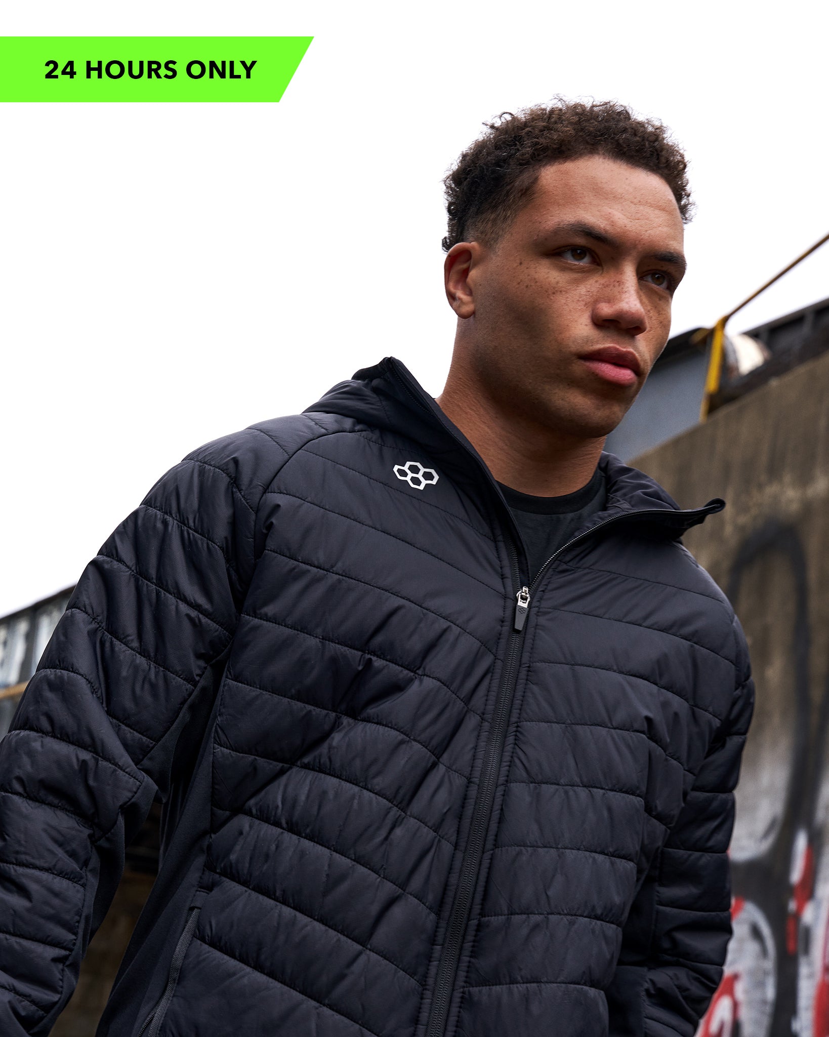 Man wearing a black quilted jacket with a brand logo, standing against an urban background.