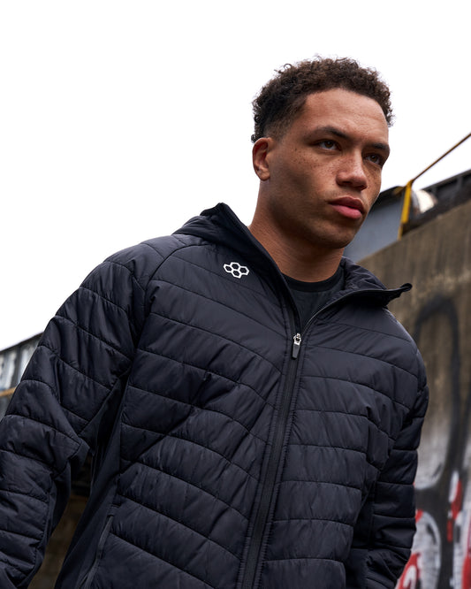 Man wearing a black quilted jacket with a brand logo, standing against an urban background.