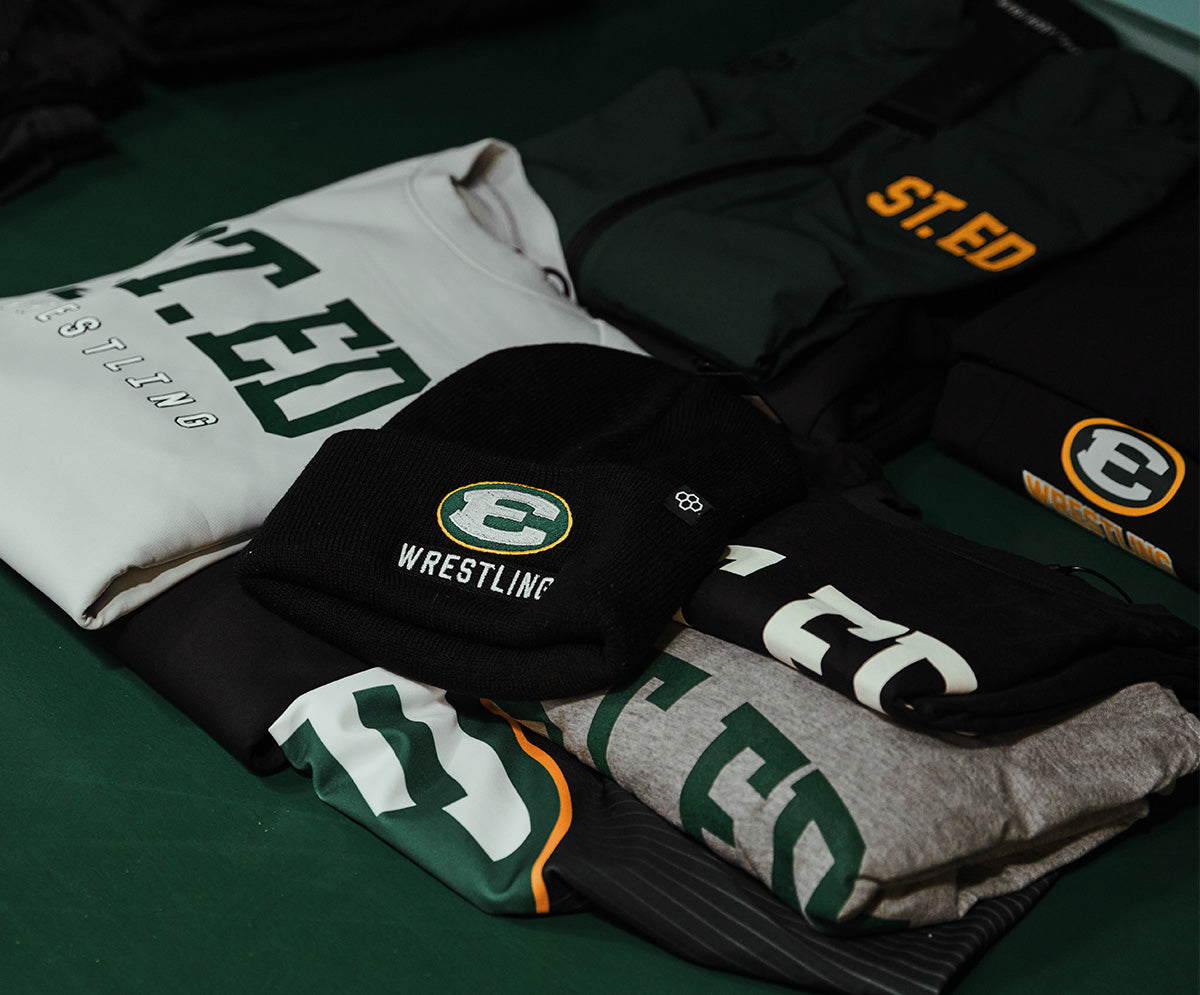 Stack of sports custom school athletic gear including hoodies and a mask with 'St. Ed' branding on a green surface.
