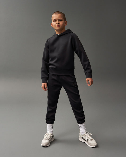A boy stands confidently in a dark tracksuit with a hood, wearing light-colored sneakers and white patterned socks against a muted gray background.