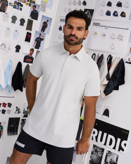 A man wearing a white short-sleeve polo shirt and black shorts, standing in front of a wall covered with fashion sketches and apparel photos. The brand name RUDIS is visible on the wall and on the shorts.