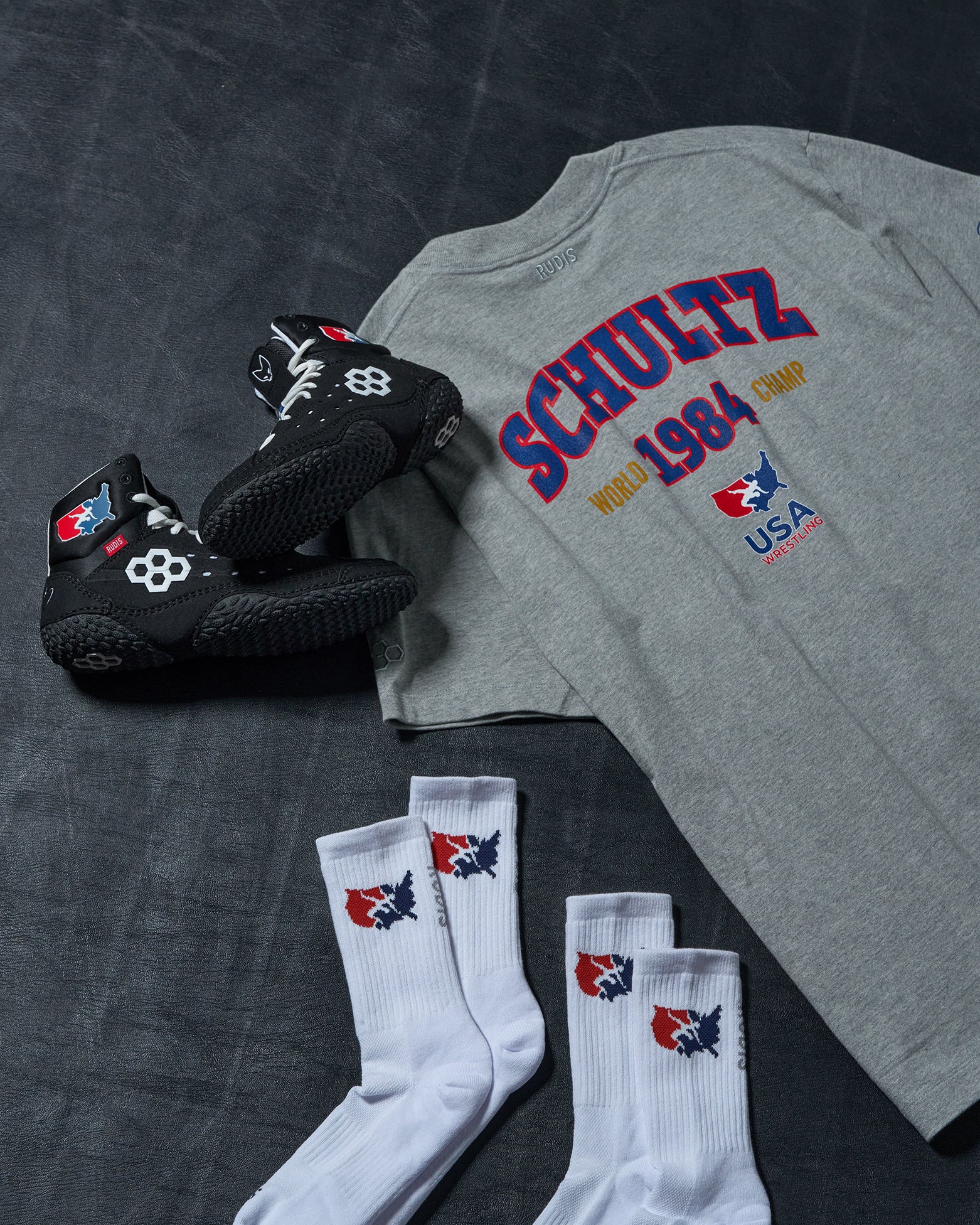 Gray t-shirt with 'Schultz 1984' design, black wrestling shoes, and white socks on a dark surface