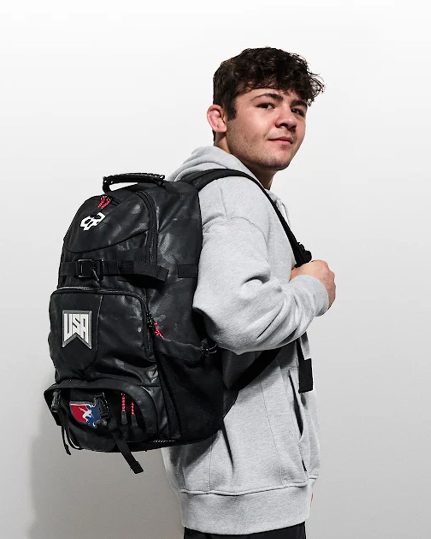 A man wearing a light gray hoodie and black pants is shown from the back, wearing a large black backpack with white and red graphics. The photo is taken against a plain light gray wall.
