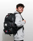 A man wearing a light gray hoodie and black pants is shown from the back, wearing a large black backpack with white and red graphics. The photo is taken against a plain light gray wall.