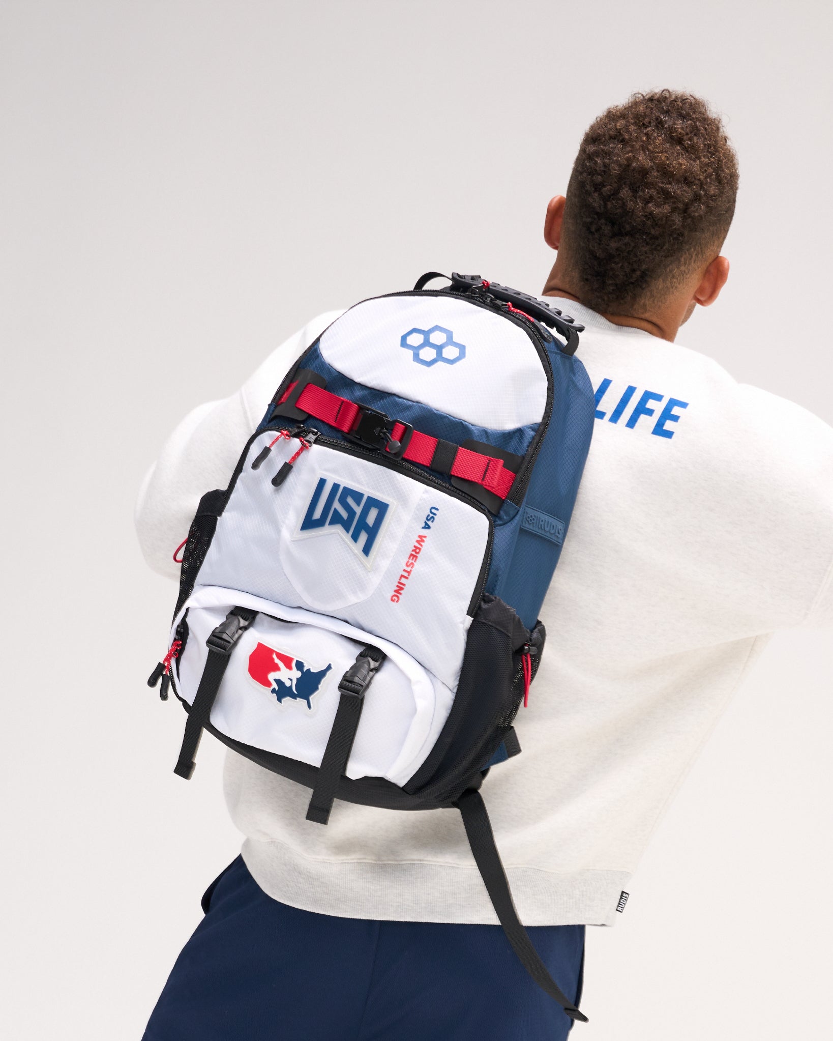Rear view of a model wearing the RUDIS USA Wrestling Genesis Gearpack III backpack, featuring red, white, and blue panels, USA Wrestling graphics, multiple zippered compartments, and adjustable straps.
