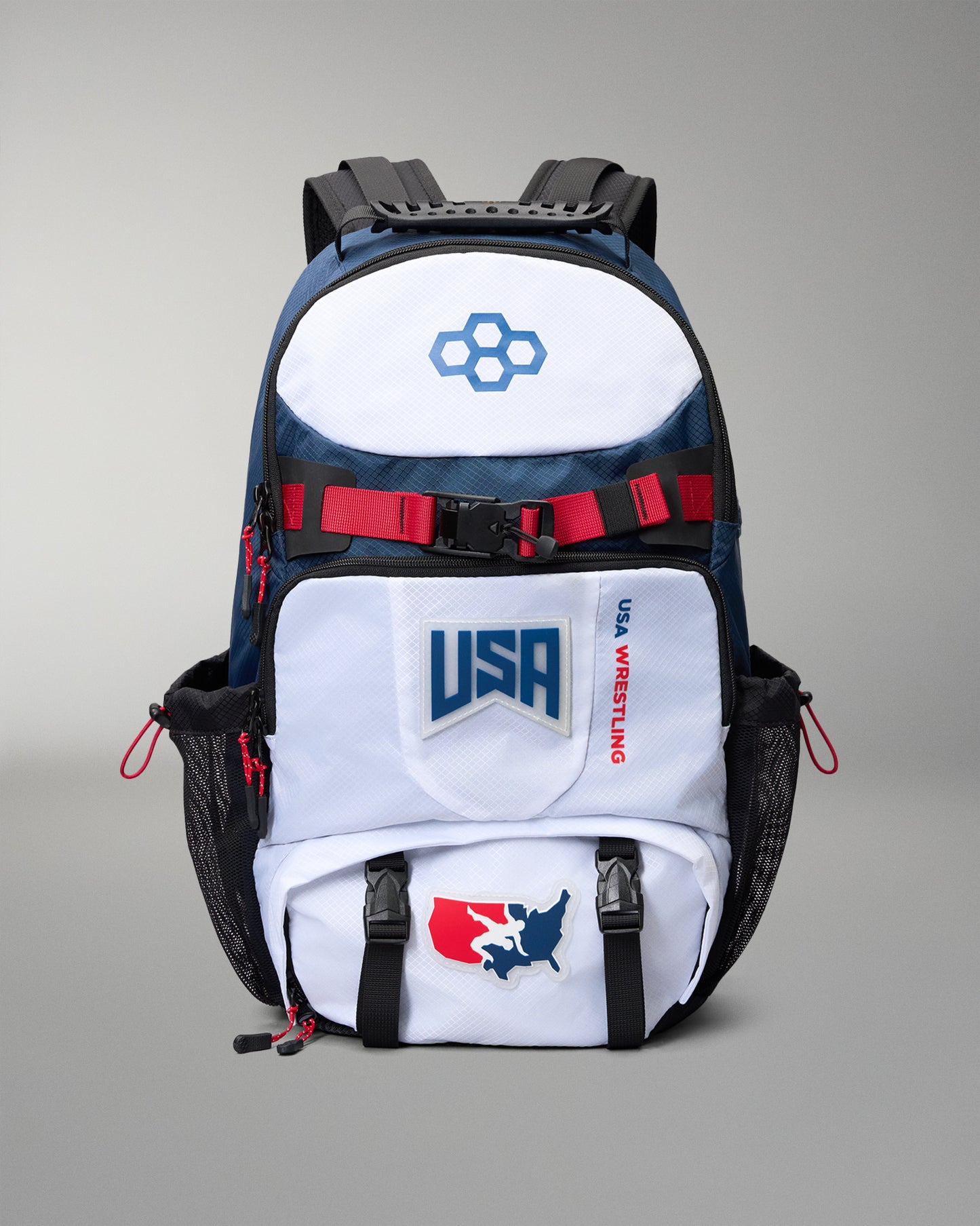 A USA Wrestling branded backpack in navy blue, white, and black, featuring red accents and team logos, shown against a neutral gray background.