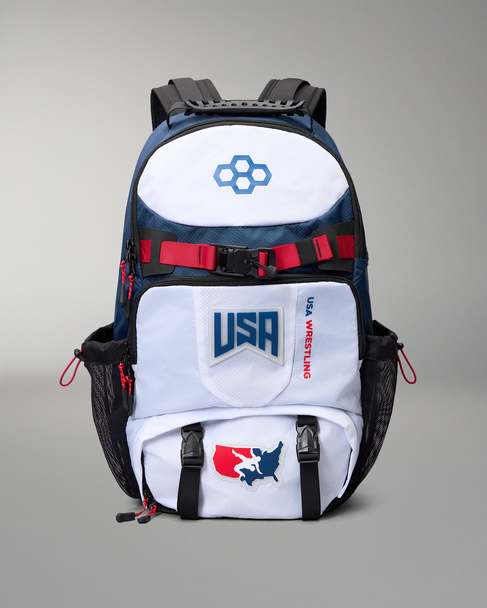A USA Wrestling branded backpack in navy blue, white, and black, featuring red accents and team logos, shown against a neutral gray background.