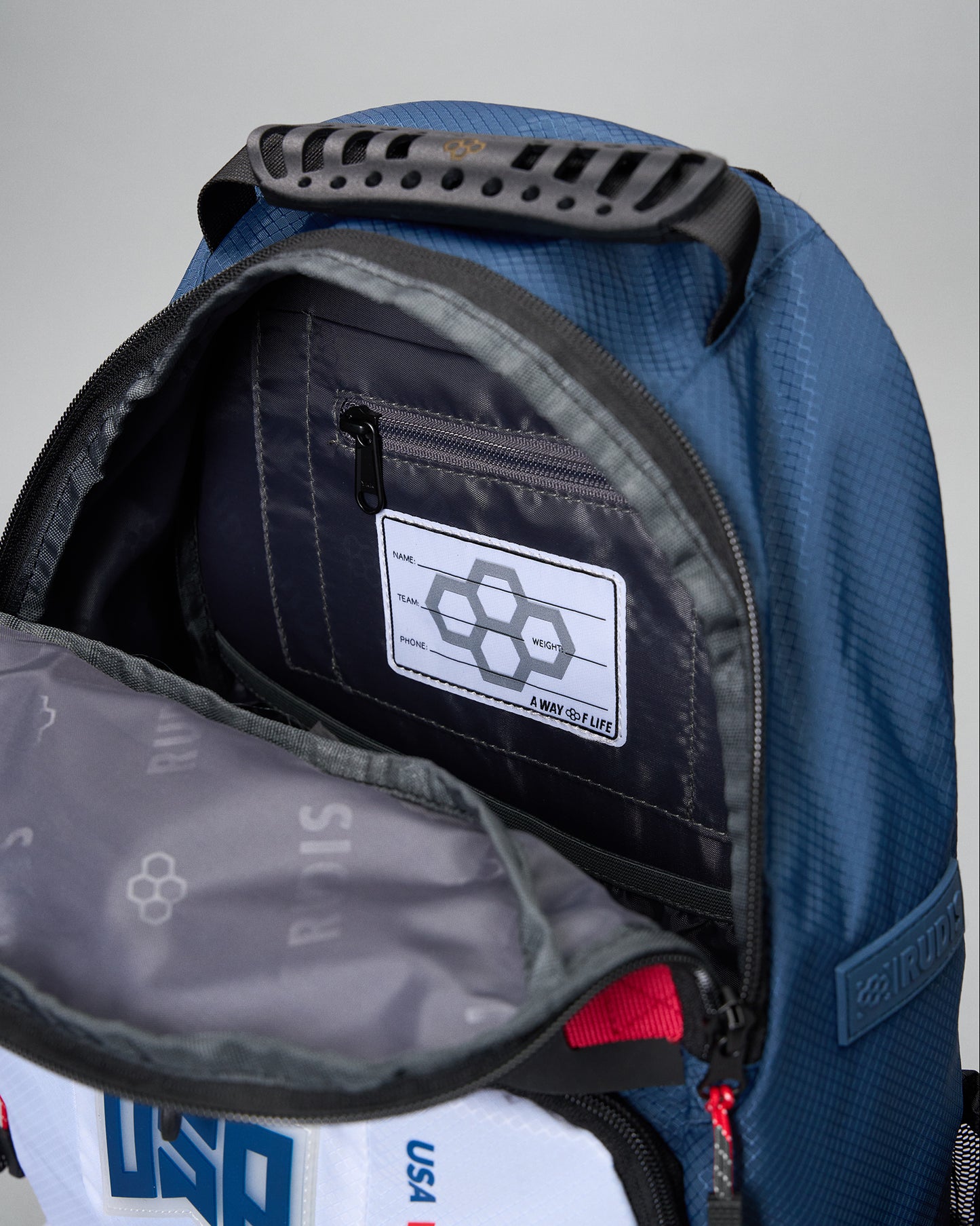 Close-up of an open blue backpack showing the interior, a zippered pocket with a white patch, and a gray inner lining. The brand name RUDIS is visible on the pocket patch and side tab.
