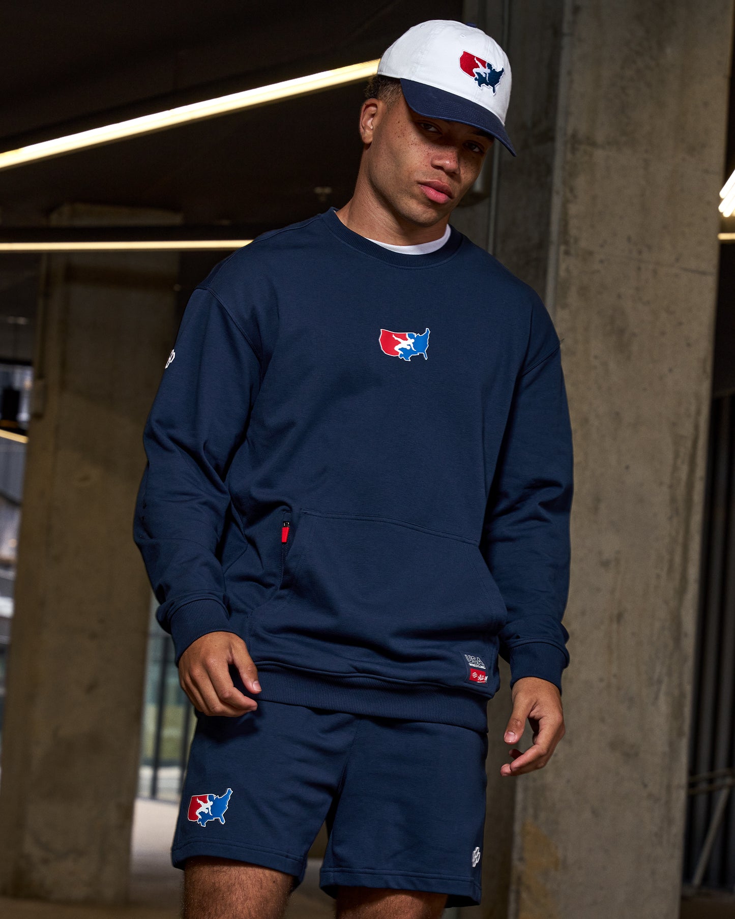 Person wearing navy blue sweatshirt and shorts with a logo, standing against a concrete wall.
