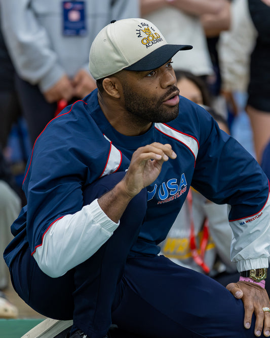 A man wearing a navy blue and white USA track and field tracksuit with 'USA ATHLETICS' on the chest, a matching baseball cap, and a gold watch, sitting in a crowd.