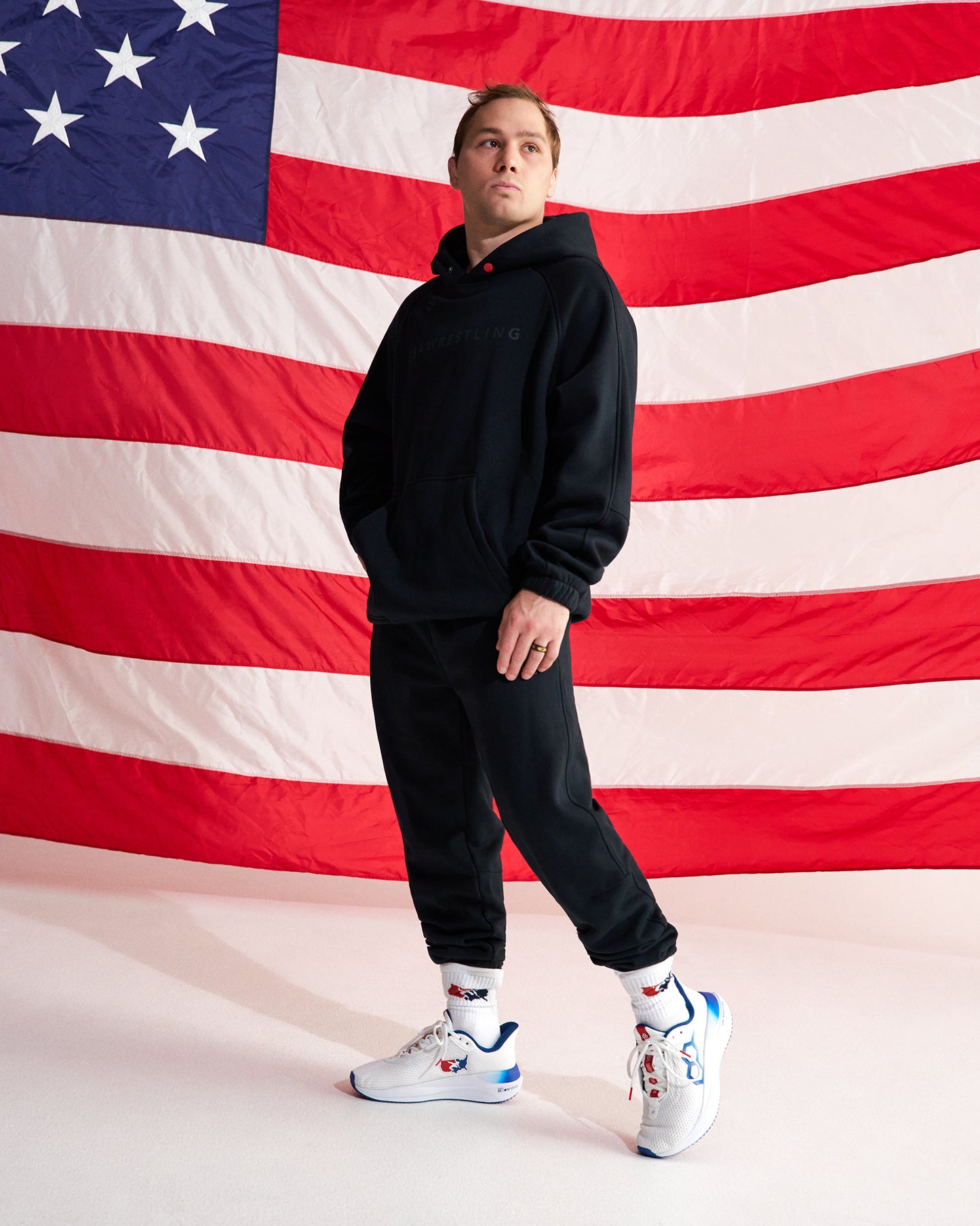 A man modeling a black hoodie and black jogger pants in front of a large American flag backdrop, wearing white sneakers with red and blue accents.