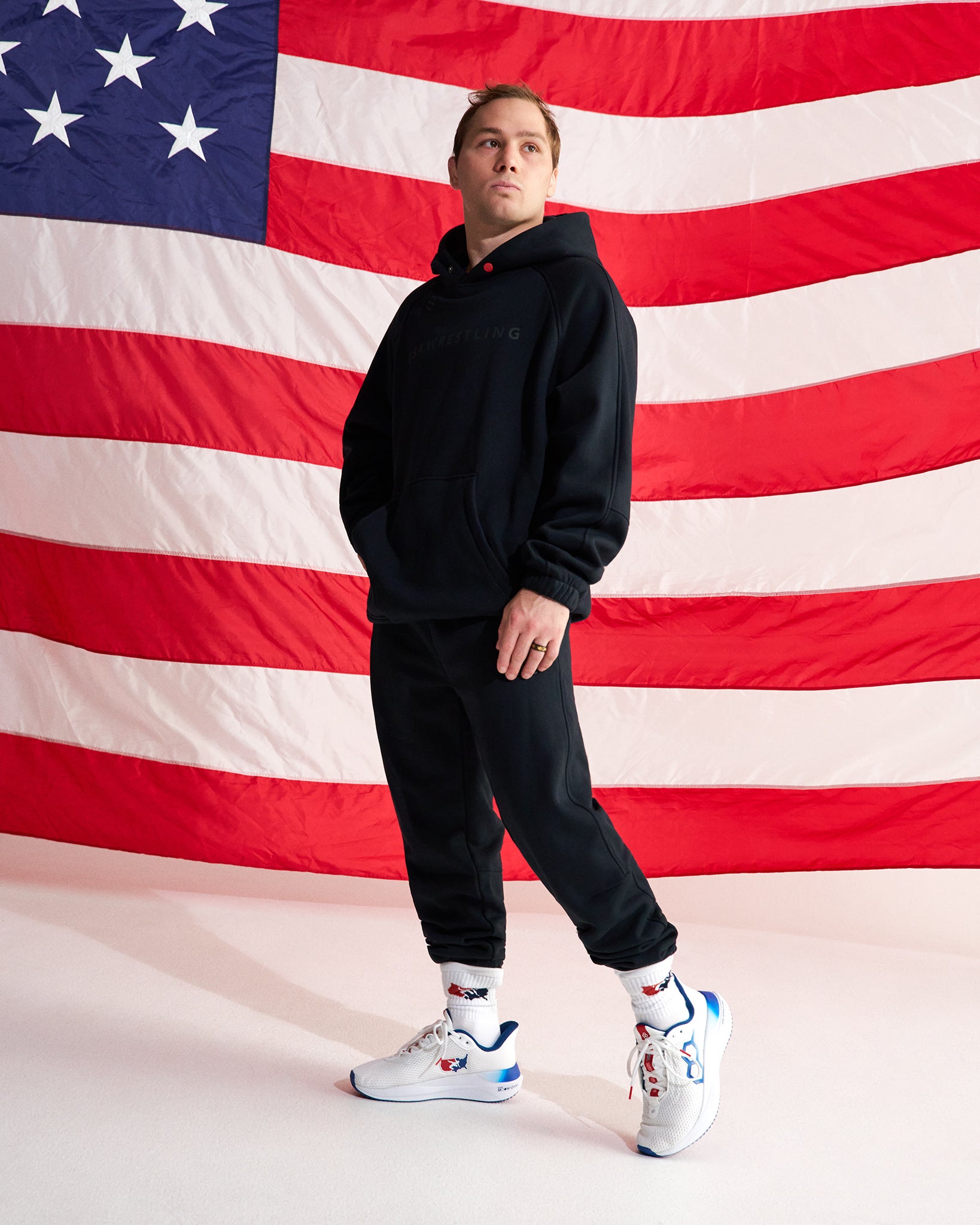 A man modeling a black hoodie and black jogger pants in front of a large American flag backdrop, wearing white sneakers with red and blue accents.