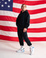 A man modeling a black hoodie and black jogger pants in front of a large American flag backdrop, wearing white sneakers with red and blue accents.