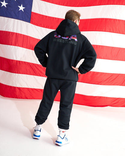 A person standing with their back to the camera, wearing a black hoodie and black sweatpants, with a large American flag backdrop behind them. The hoodie features a graphic on the back with the word 'WRESTLING' and a small American flag design.