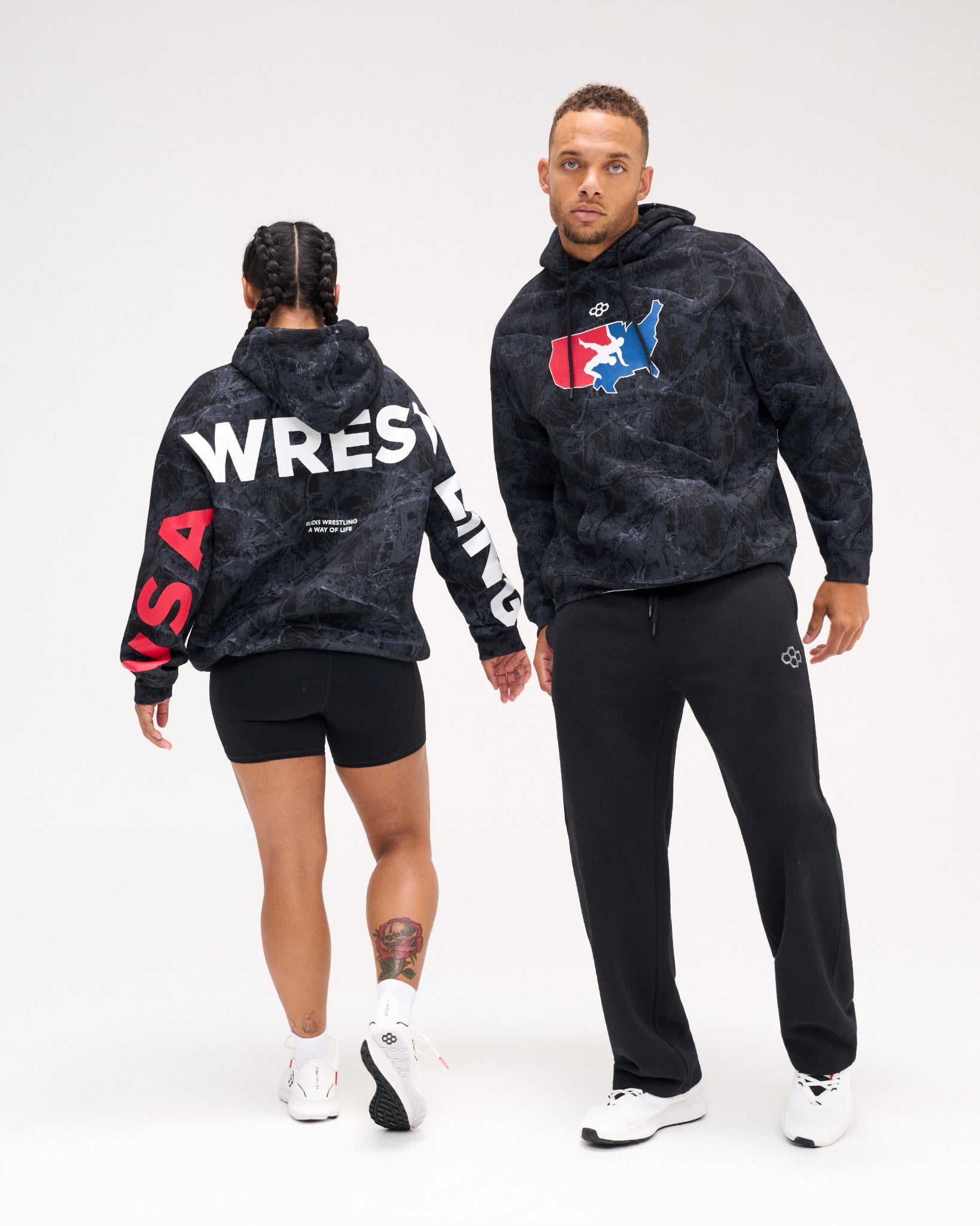 Male and female models wearing the RUDIS USA Wrestling Map Relaxed Comfort Fleece Hoodie in Black Camo, showing the USA map chest logo and bold WRESTLING back and sleeve graphics.