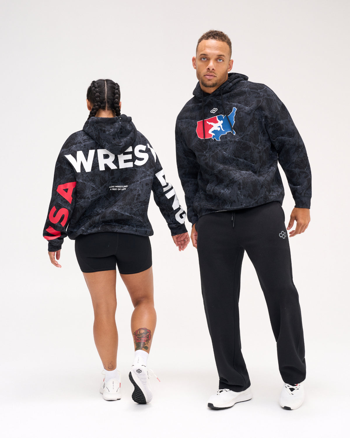 Male and female models wearing the RUDIS USA Wrestling Map Relaxed Comfort Fleece Hoodie in Black Camo, showing the USA map chest logo and bold WRESTLING back and sleeve graphics.