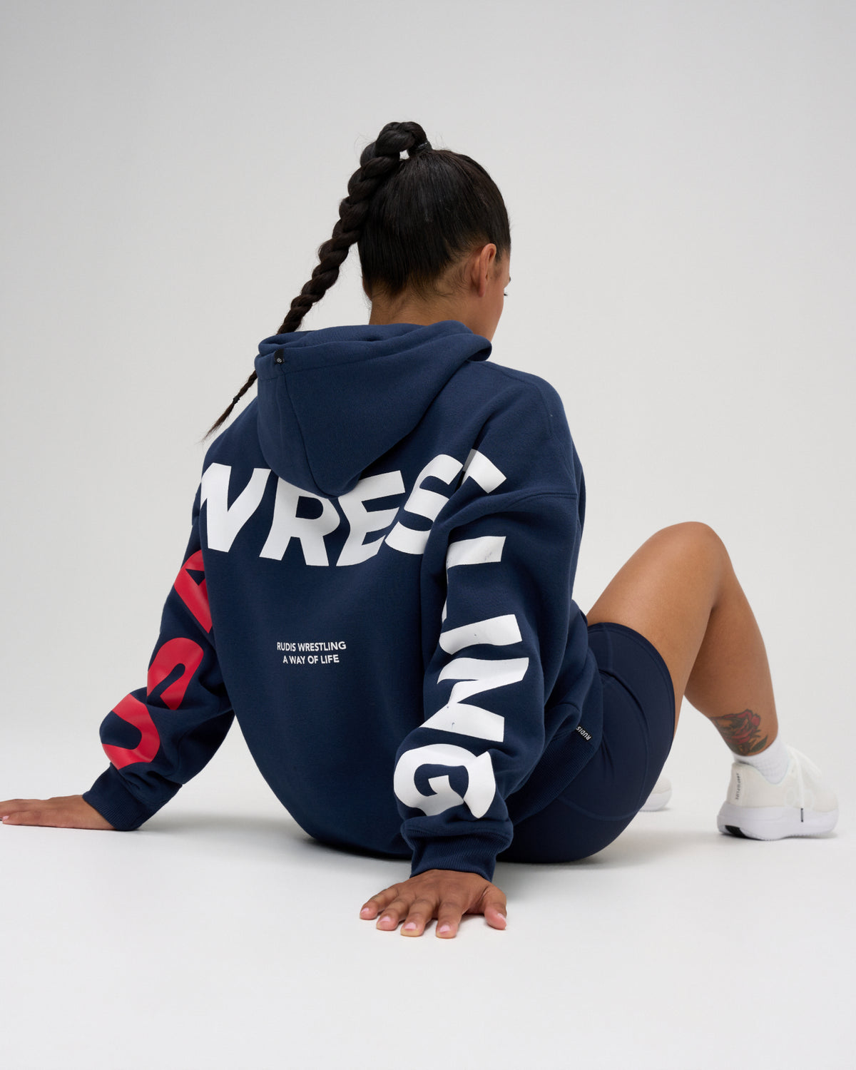 Rear view of female model seated in the RUDIS USA Wrestling Map fleece hoodie in Navy, featuring oversized WRESTLING lettering and RUDIS Wrestling A Way of Life back detail.