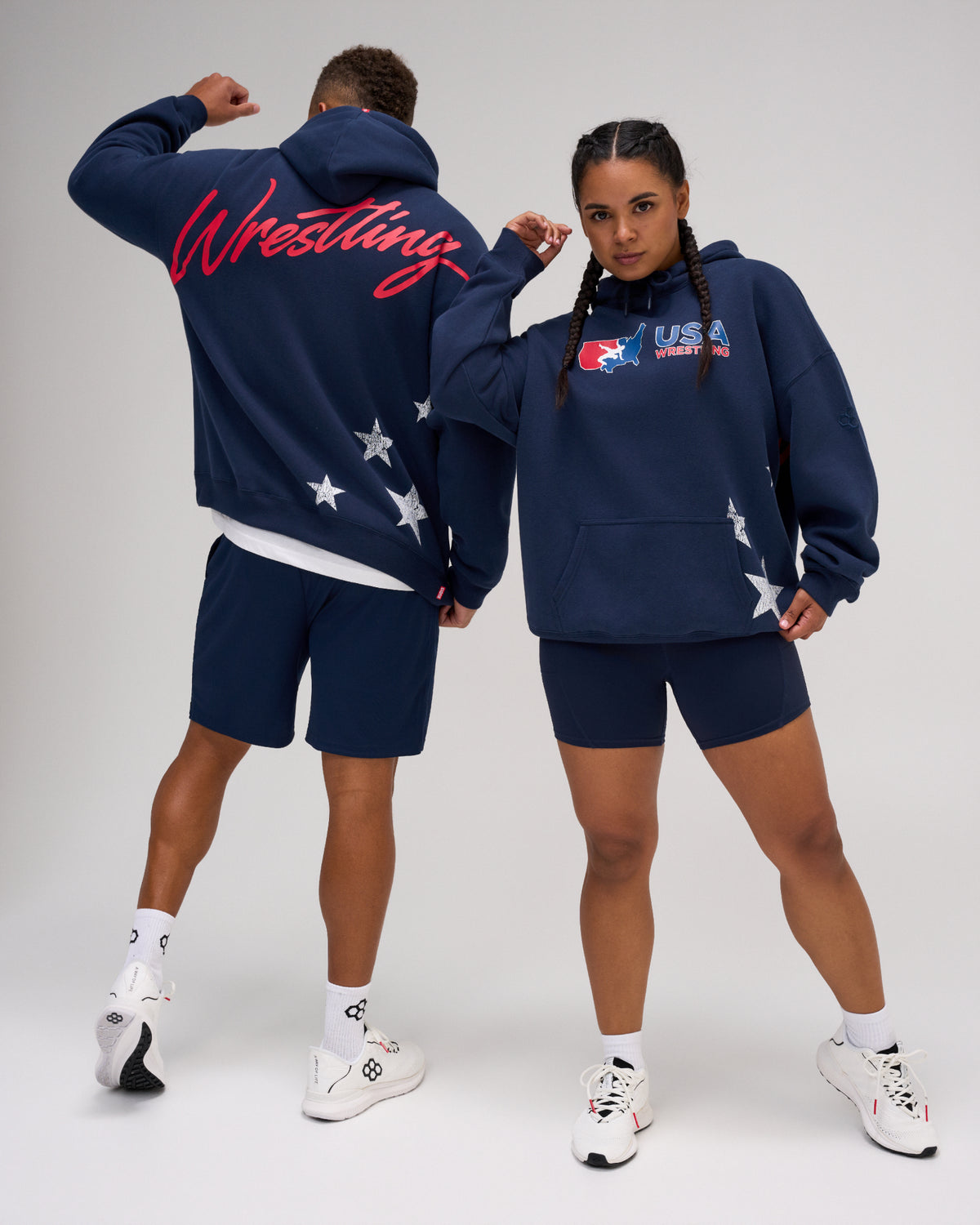 Male and female models wearing the RUDIS USA Wrestling Script Relaxed Comfort Fleece Hoodie in Navy, showing the USA Wrestling chest logo, red Wrestling back script and white star graphics.