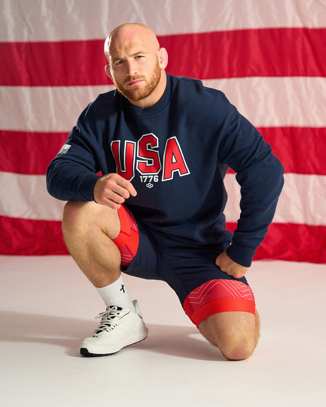 Man wearing a navy blue 'USA 1776' sweatshirt with red and white shorts in front of an American flag.