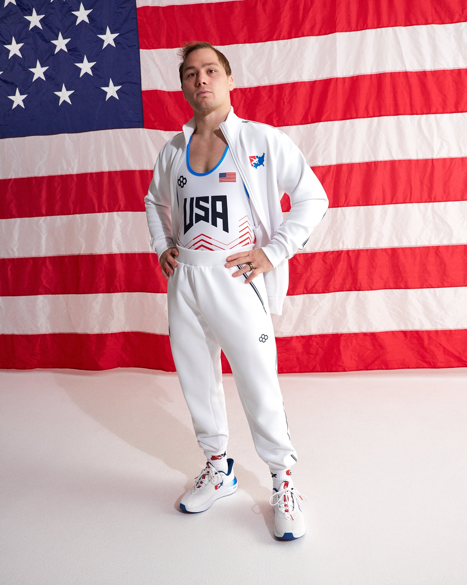 A man modeling a white USA track and field uniform set consisting of a zip-up jacket with 'USA' printed on the chest and matching sweatpants, posed in front of a large American flag backdrop.