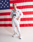 A man modeling a white USA track and field uniform set consisting of a zip-up jacket with 'USA' printed on the chest and matching sweatpants, posed in front of a large American flag backdrop.