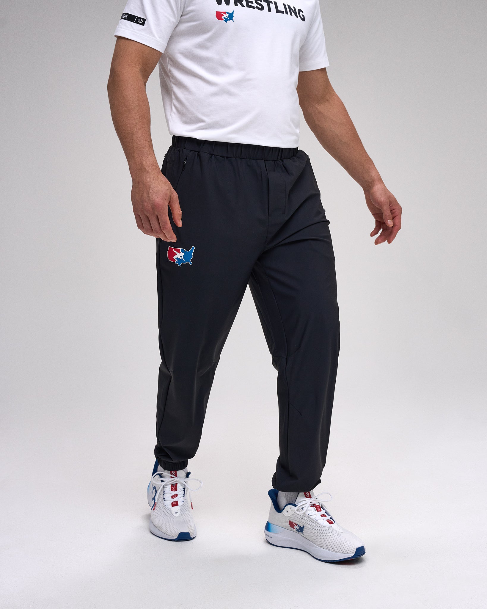 Person wearing a white t-shirt with 'Wrestling' text, black pants with a 'USA Wrestling' logo, and white sneakers on a gray background