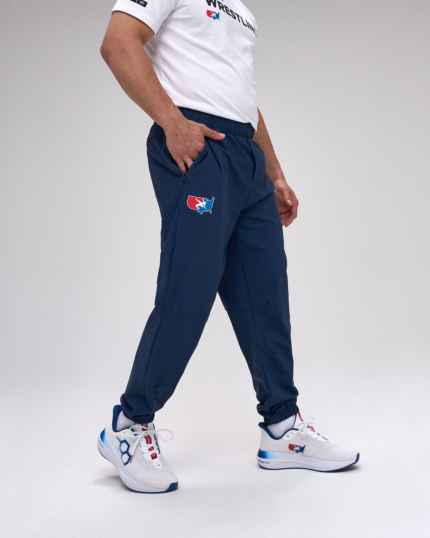 Person wearing navy tracksuit pants with a 'USA Wrestling' logo and white sneakers on a light gray background