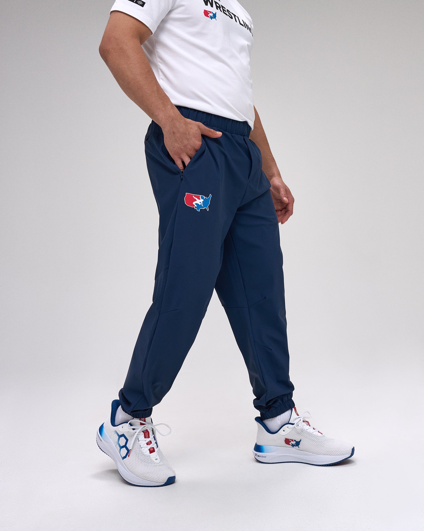 Person wearing navy tracksuit pants with a 'USA Wrestling' logo and white sneakers on a light gray background