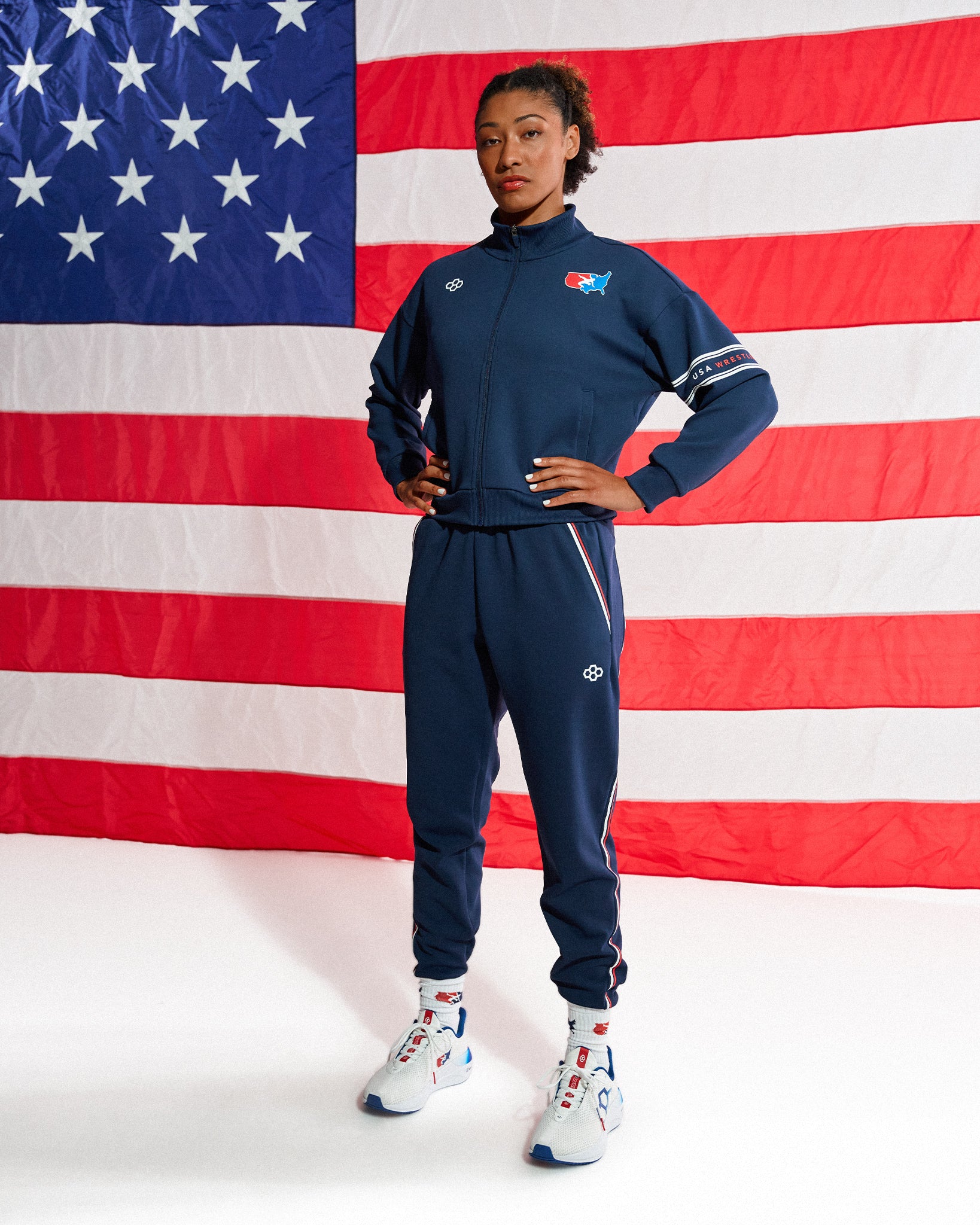 A model wearing a navy blue athletic tracksuit with white and red accents, standing in front of a large American flag backdrop. The tracksuit features a zip-up jacket and matching jogger pants with white side stripes and logos.