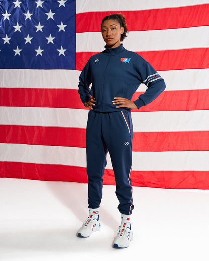 A model wearing a navy blue athletic tracksuit with white and red accents, standing in front of a large American flag backdrop. The tracksuit features a zip-up jacket and matching jogger pants with white side stripes and logos.