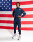 A model wearing a navy blue athletic tracksuit with white and red accents, standing in front of a large American flag backdrop. The tracksuit features a zip-up jacket and matching jogger pants with white side stripes and logos.