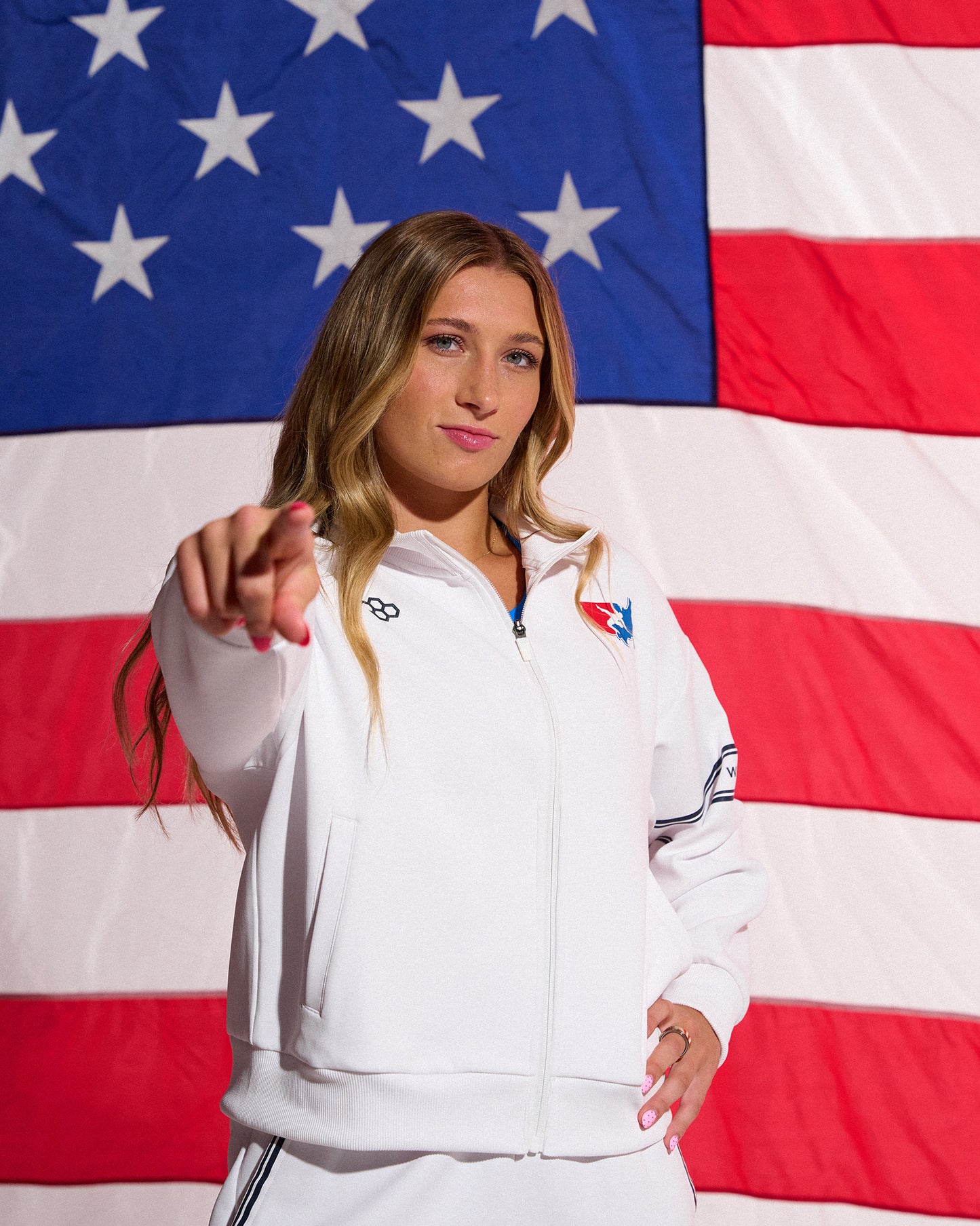 Female athlete wearing the white USA Wrestling Chronicle Elite Jacket by RUDIS points toward the camera while standing in front of a large American flag, showcasing official USA Wrestling apparel.