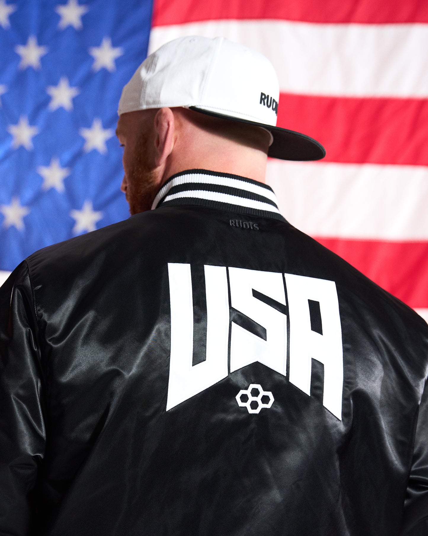 Person wearing a black jacket with 'USA' on the back in front of an American flag