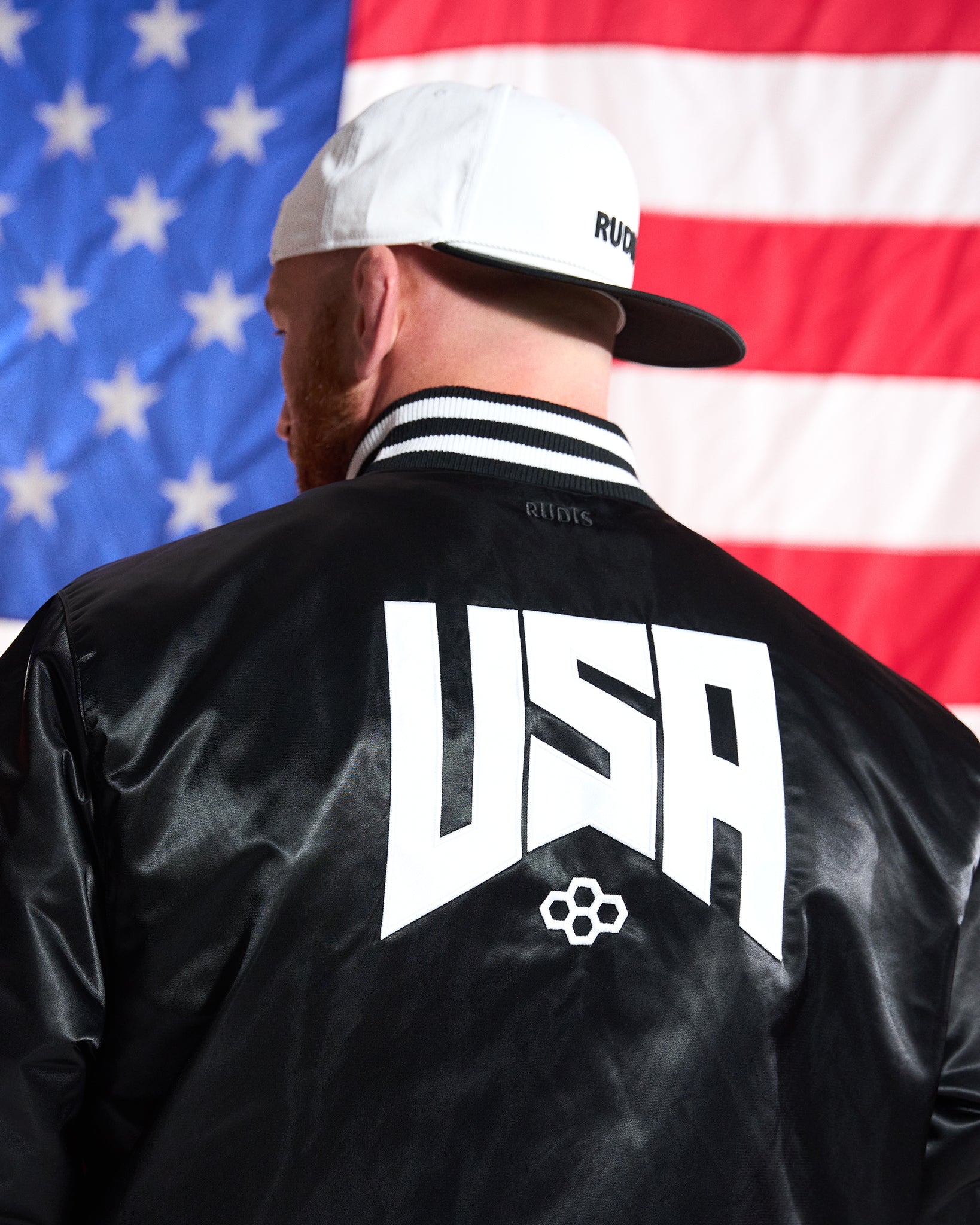 Person wearing a black jacket with 'USA' on the back in front of an American flag