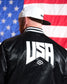 Person wearing a black jacket with 'USA' on the back in front of an American flag