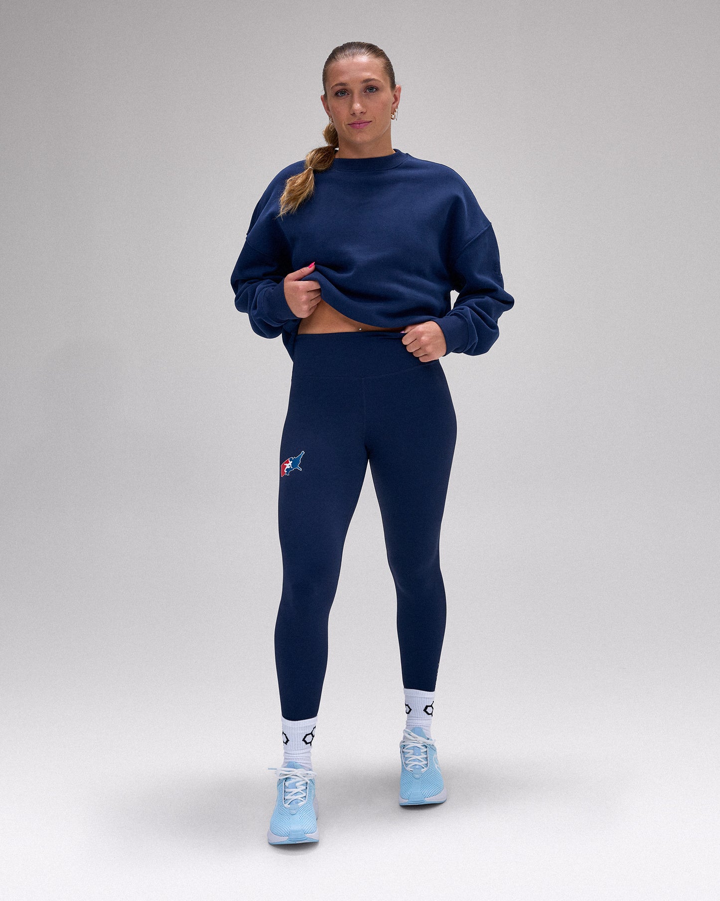 A woman modeling a matching navy blue activewear set consisting of a long-sleeve sweatshirt and high-waisted leggings, paired with white socks and light blue sneakers, against a neutral gray studio background.