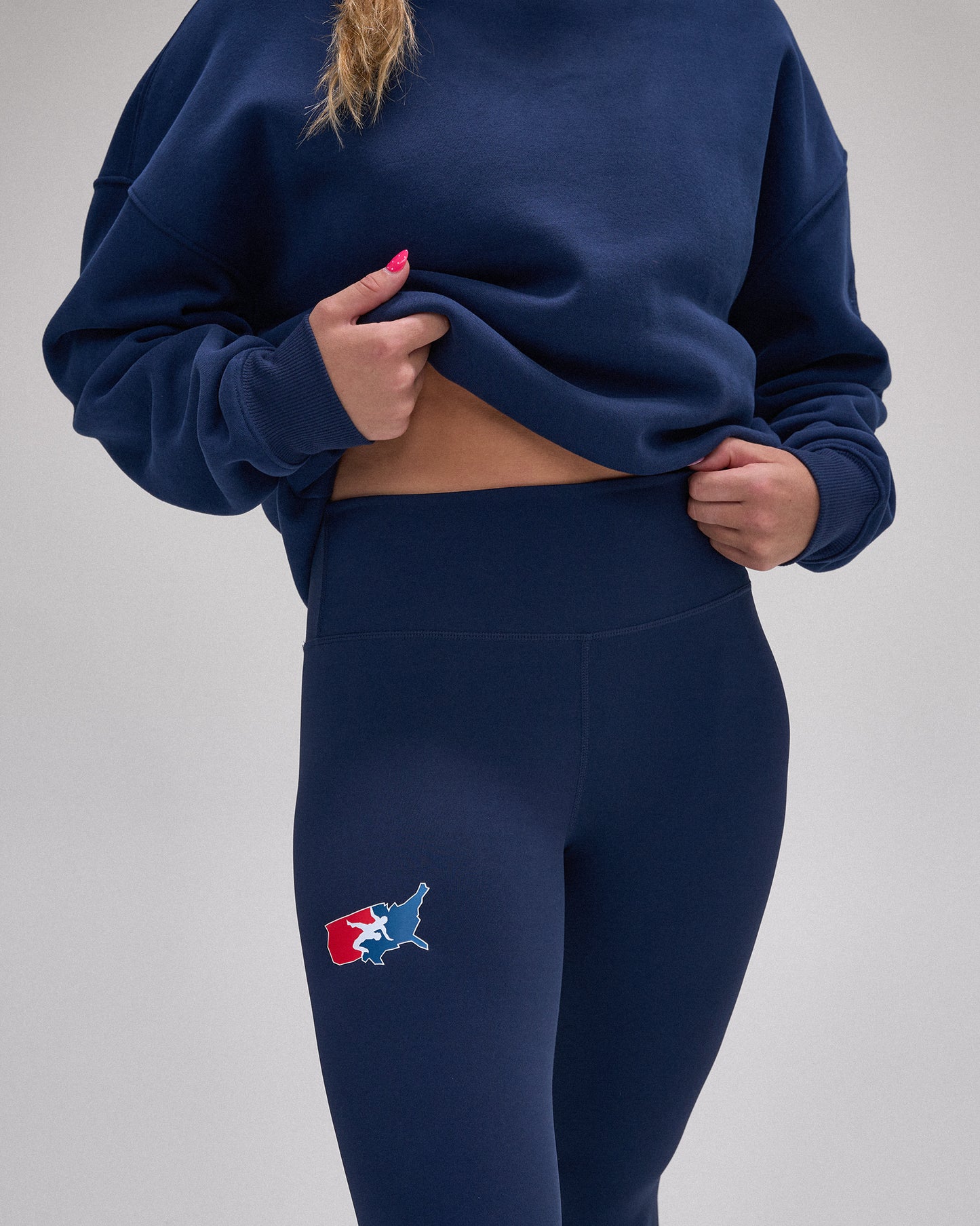 A person wearing a navy blue sweatshirt and matching high-waisted leggings, lifting the sweatshirt to show the midriff. The leggings feature a small embroidered Texas state map logo in red, white, and blue on the left thigh, set against a plain light gray studio background.
