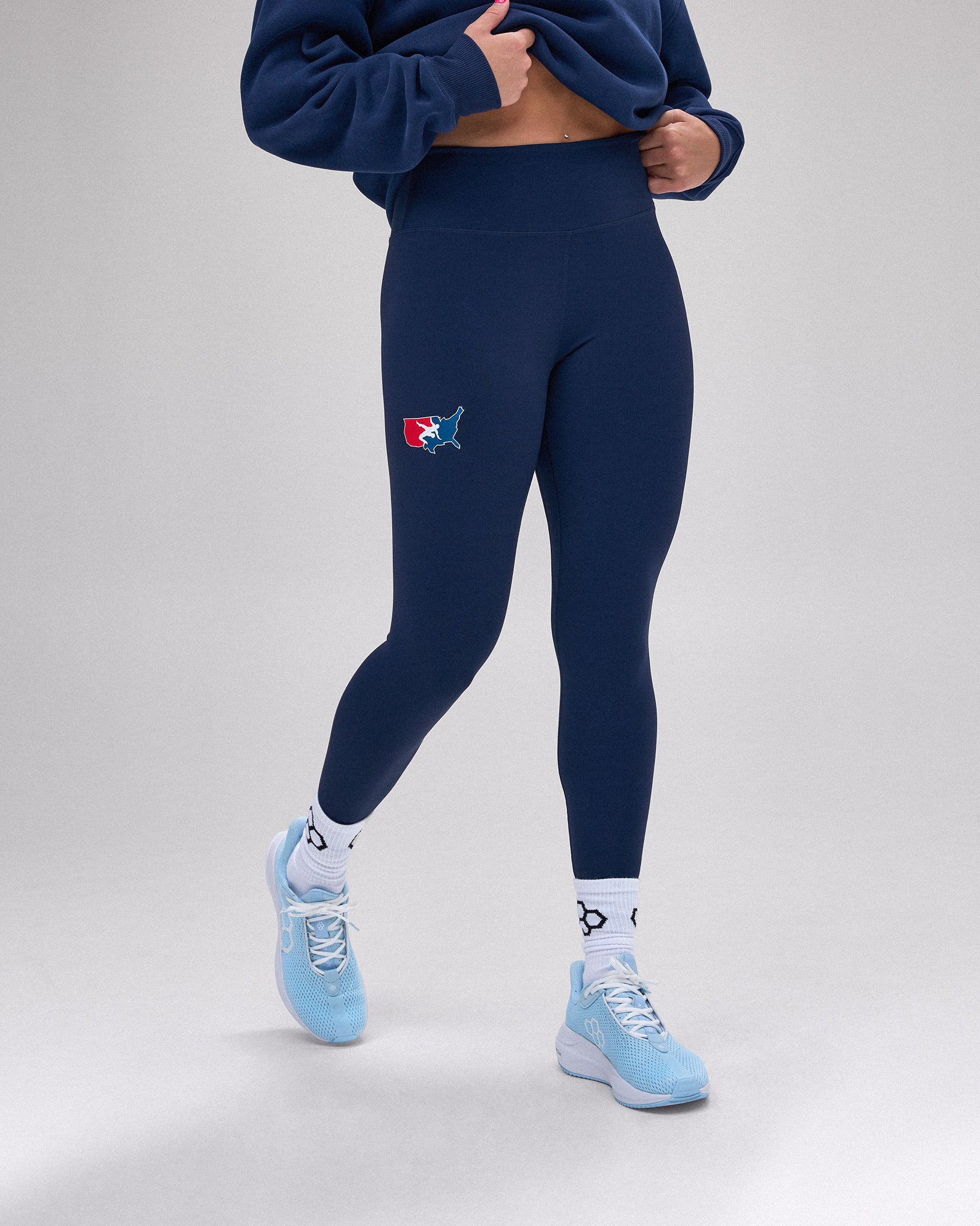 A model wearing navy blue high-waisted leggings with a small red and blue shark logo on the left thigh, paired with a navy cropped sweatshirt, white socks, and light blue sneakers, against a light gray studio background.