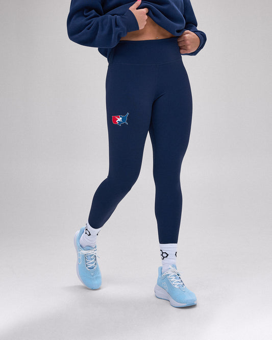 A model wearing navy blue high-waisted leggings with a small red and blue shark logo on the left thigh, paired with a navy cropped sweatshirt, white socks, and light blue sneakers, against a light gray studio background.