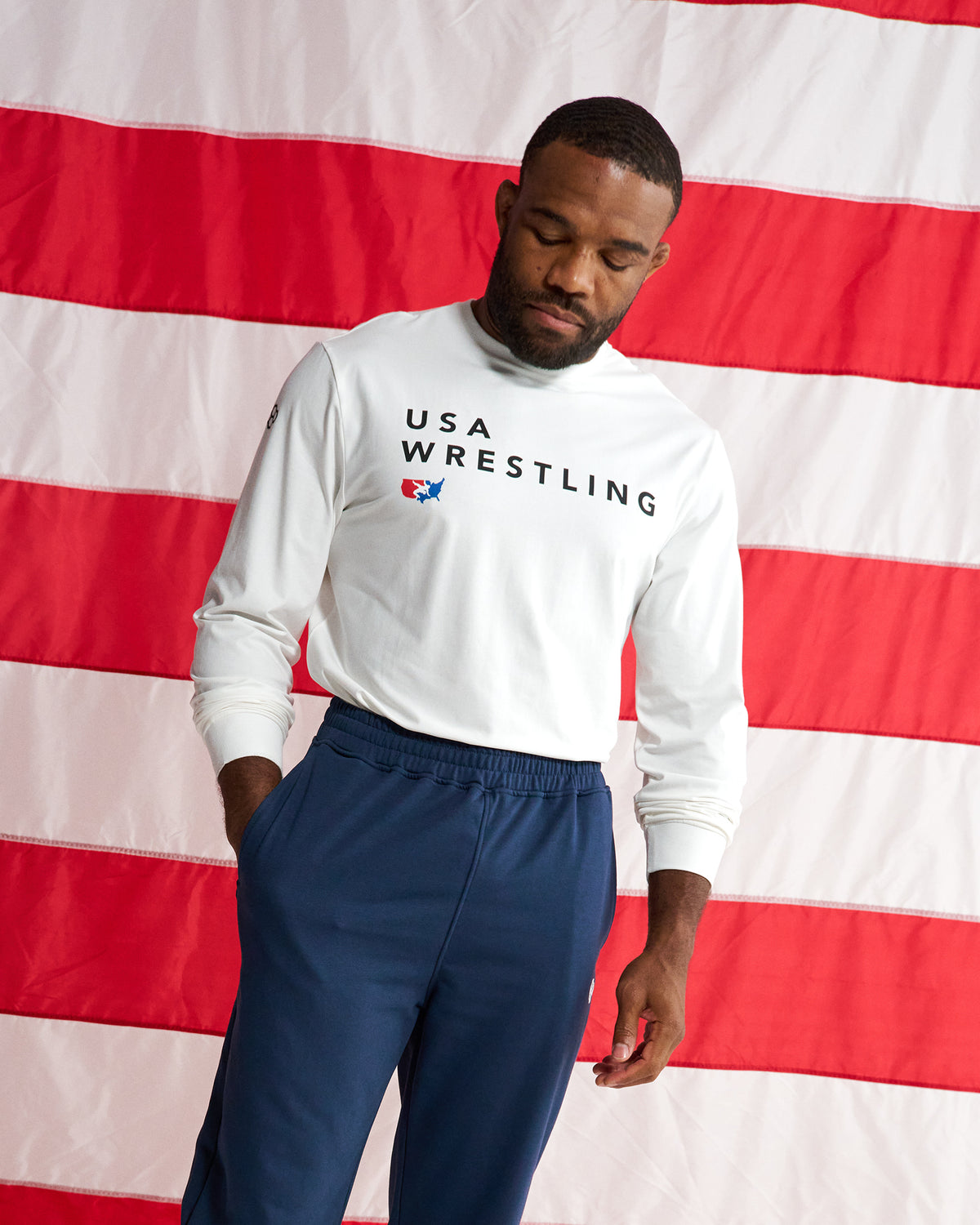 A man wearing a white long-sleeve turtleneck sweatshirt with 'USA WRESTLING' printed in black across the chest, paired with navy blue sweatpants, standing in front of a large red and white striped flag backdrop.