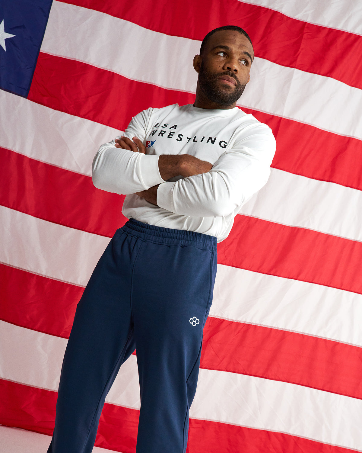 A man stands with arms crossed in front of a large American flag backdrop, wearing a white long-sleeve shirt with 'USA WRESTLING' printed across the chest and navy blue sweatpants.
