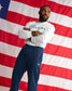 A man stands with arms crossed in front of a large American flag backdrop, wearing a white long-sleeve shirt with 'USA WRESTLING' printed across the chest and navy blue sweatpants.