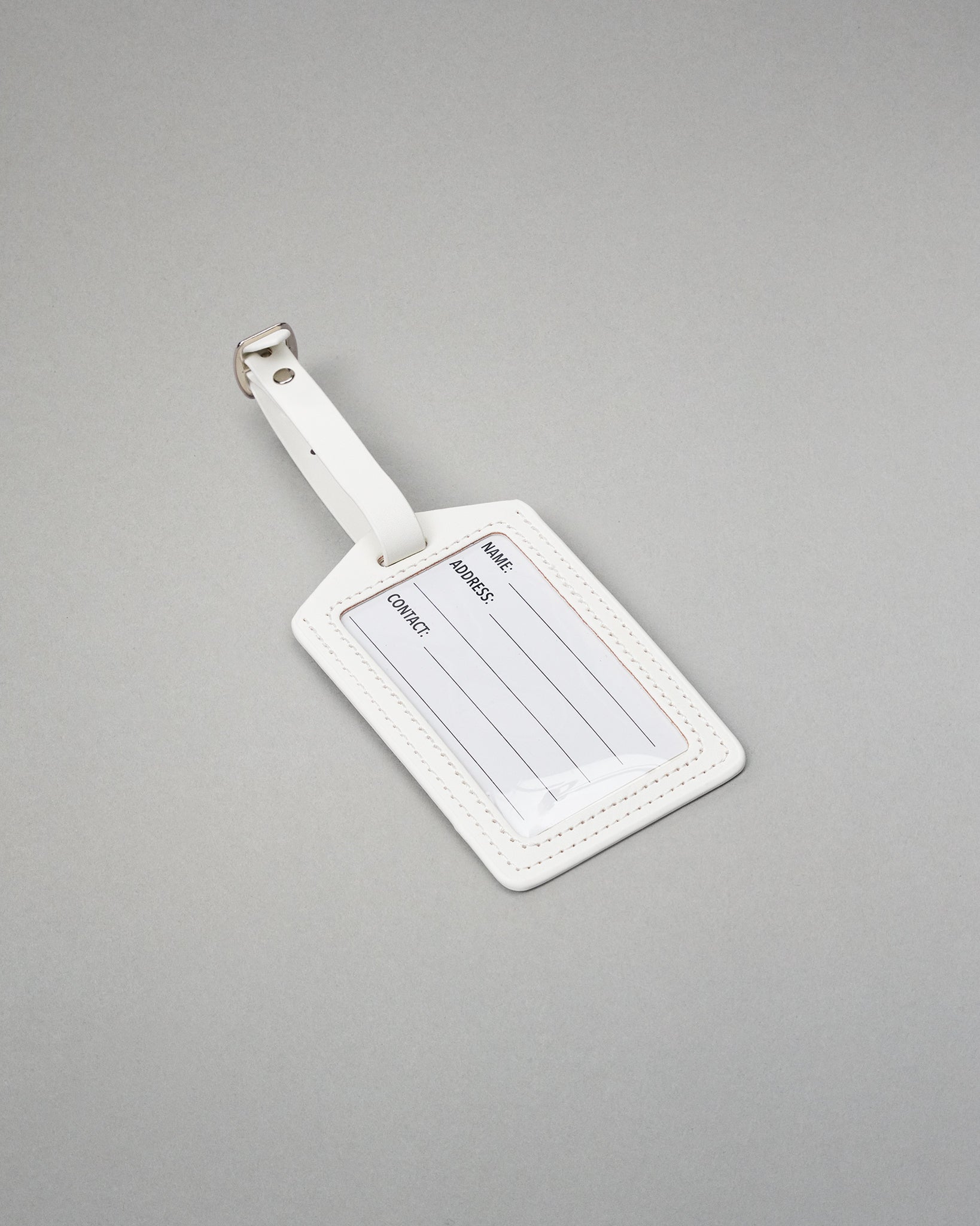 A white leather luggage tag with a silver metal buckle, featuring blank lines for name, address, and contact information, set against a solid gray background.