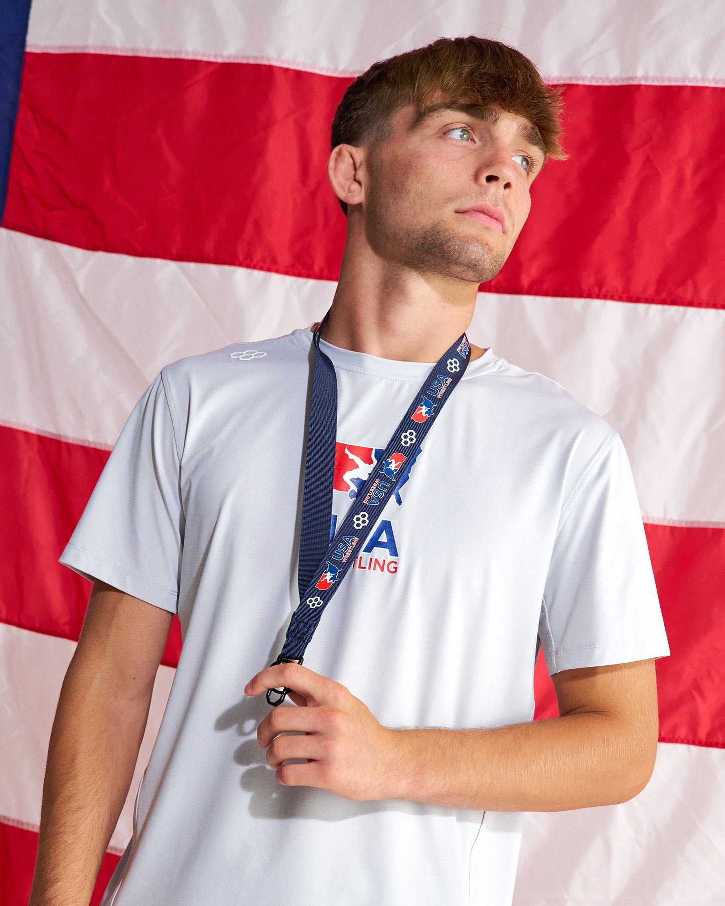 A young man wearing a white USA Wrestling t-shirt and a navy blue lanyard with the USA National Team logo, posed in front of a large American flag.